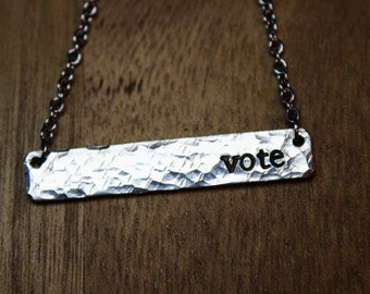 以下が含まれることがあります： 黒い文字で「vote」と書かれたハンマーで叩かれたバーペンダントが付いたシルバーネックレス