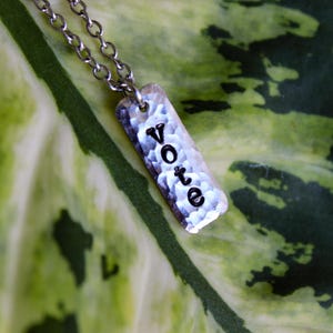 May include: A silver-colored pendant necklace with a rectangular charm that reads "vote" in black letters. The necklace hangs against a backdrop of a green and yellow leaf.