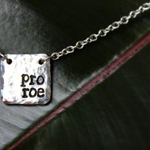 Puede incluir: Collar plateado con un colgante cuadrado. El colgante tiene una textura martillada y las palabras "pro roe" en letras negras. La cadena es delicada y el fondo es una hoja verde oscuro.