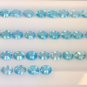 May include: A collection of oval-shaped, light blue gemstones arranged in rows on a white surface.