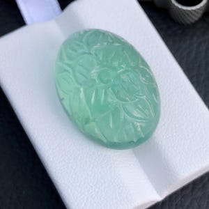 May include: A carved green gemstone with a floral design. The gemstone is oval shaped and is displayed on a white background.
