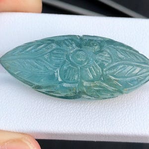 May include: A carved aquamarine gemstone pendant in the shape of a leaf with a flower design. The pendant is a light blue color and has a smooth, polished finish.