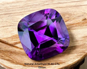 Könnte beinhalten: Ein facettierter, kissenförmiger natürlicher Amethyst-Edelstein mit tiefvioletten Farbtönen. Der Edelstein liegt auf einer Holzoberfläche. Text auf dem Bild: Natural Amethyst 10.40 CTs. 13mm.