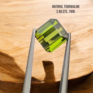 May include: A green tourmaline gemstone, weighing 2.80 carats and measuring 7mm, is held by tweezers against a wooden background. NATURAL TOURMALINE 2.80 CTS. 7MM. is written above the gemstone.