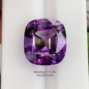 May include: A cushion-cut amethyst gemstone, displaying a deep purple hue. The gem is 16x15x11mm and weighs 17 carats. The facets reflect light, creating a brilliant display. The text "Amethyst 17 CTs. 16x15x11mm." is visible.