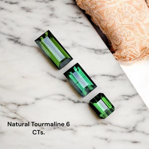 May include: Three rectangular, faceted green tourmaline gemstones are displayed on a white marble surface. The gemstones are arranged diagonally, with the largest at the top and the smallest at the bottom. The text "Natural Tourmaline 6 CTs." is at the bottom.