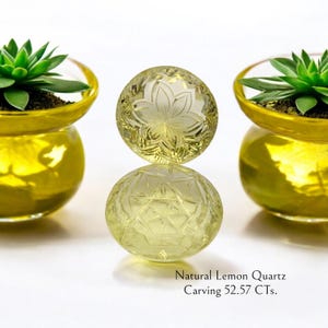 May include: Two yellow-green quartz spheres, one with a carved floral design, are stacked. The bottom sphere has a geometric pattern. The text reads "Natural Lemon Quartz Carving 52.57 CTs." Two small succulent plants in gold-colored glass pots flank the spheres.