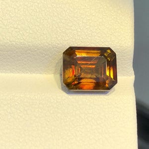 May include: A rectangular, faceted gemstone with a deep orange and brown color. The gemstone has a slightly reddish hue and is set on a white background.