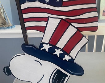May include: A wooden cutout of Snoopy dressed as Uncle Sam, wearing a blue top hat with white stars and a red, white, and blue scarf. He is holding a small American flag in his right hand. The cutout is standing on a wooden easel.