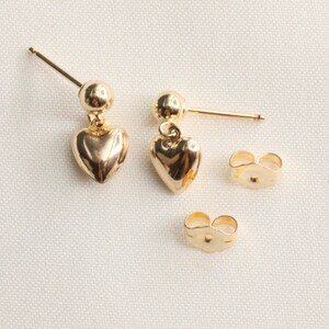 May include: A pair of gold heart dangle earrings with a small ball at the top of the post. The earrings come with two sets of backings.