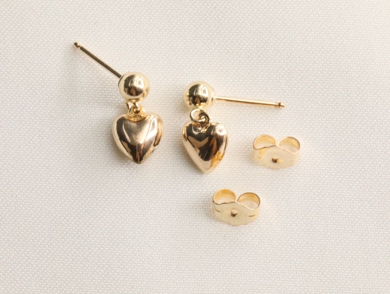 May include: A pair of gold heart dangle earrings with a small ball at the top of the post. The earrings come with two sets of backings.