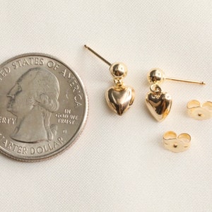 May include: A pair of gold heart-shaped dangle earrings with a small ball at the top of each earring. The earrings are shown next to a US quarter for size comparison.