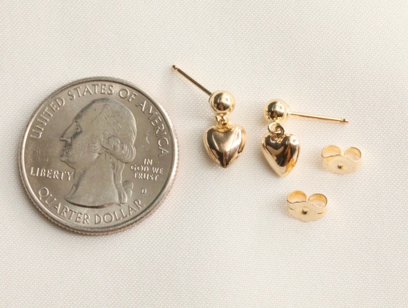 May include: A pair of gold heart-shaped dangle earrings with a small ball at the top of each earring. The earrings are shown next to a US quarter for size comparison.