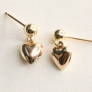 May include: A pair of gold heart-shaped dangle earrings. Each earring features a small gold ball at the top and a larger heart-shaped charm hanging below.
