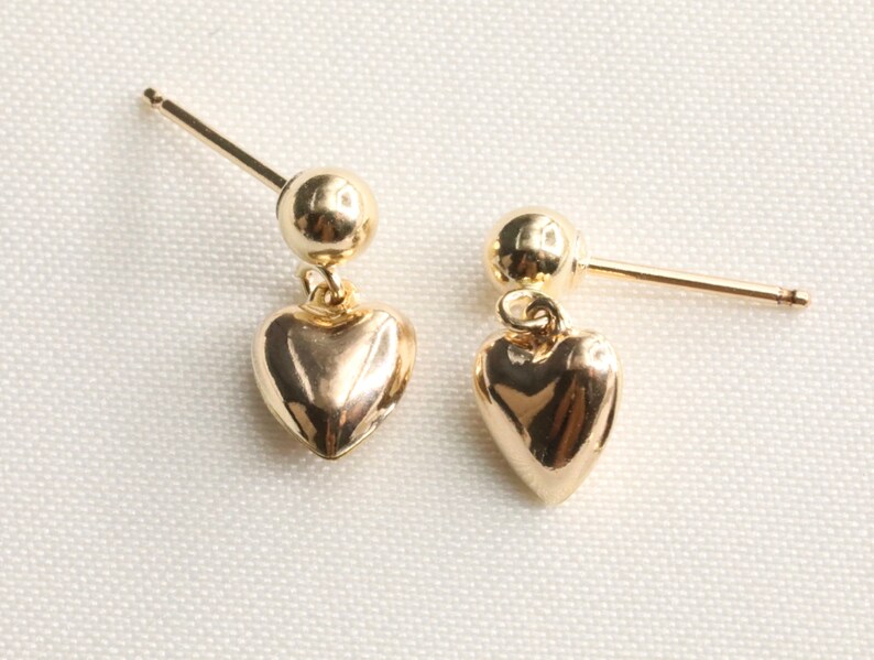 May include: A pair of gold heart-shaped dangle earrings. Each earring features a small gold ball at the top and a larger heart-shaped charm hanging below.