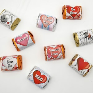 Könnte beinhalten: Eine Sammlung einzeln verpackter Süßigkeiten mit verschiedenen Valentinstagsbotschaften und Herzmotiven. Die Verpackungen sind silber, orange und goldfarben, mit Botschaften wie "Ich liebe dich", "Be Mine" und "Süße Liebe".