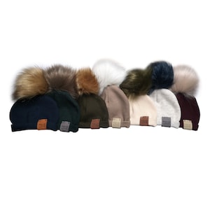 Può includere: Assortimento di dieci berretti di lana con pon pon in pelliccia sintetica. I berretti sono di vari colori, tra cui blu navy, verde scuro, verde oliva, beige, beige chiaro, grigio e bordeaux. Ogni berretto ha una piccola etichetta in pelle con le parole "Made with Love" su di essa.