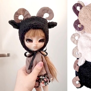 Può includere: Un cappello da ariete in peluche nero e marrone con corna ricciute. Il cappello è indossato da una bambola con lunghi capelli castani, occhi marroni e una gonna rosa a fiori.