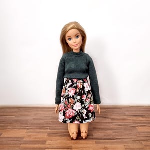 Puede incluir: Una muñeca con un suéter de manga larga verde oscuro y una falda negra con estampado floral.