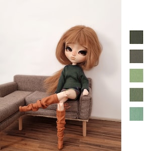 Puede incluir: Una muñeca con el pelo largo y rojo está sentada en un sofá marrón. Lleva un jersey verde, una falda de cuadros y botas marrones. La muñeca tiene ojos azules y una cara pintada.