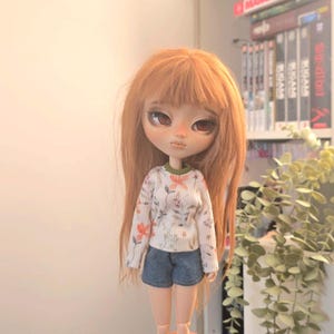 Peut inclure: Une poupée aux longs cheveux roux, vêtue d'un pull blanc à motifs floraux et d'un short en jean. La poupée présente des traits de visage détaillés et est posée devant un fond neutre.