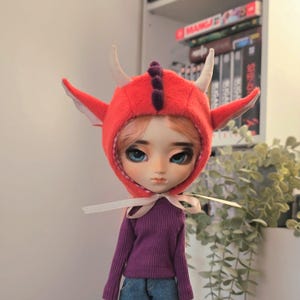 Può includere: Una bambola con un cappuccio da drago rosso con corna bianche e lembi a forma di orecchio. La bambola ha occhi blu, capelli rosa e un maglione viola. Un nastro bianco è legato intorno al collo. È un oggetto da collezione a tema fantasy.