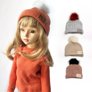 May include: A doll wearing a coral beanie with a white pom pom and the text "Made with LOVE". Three additional beanies are shown in the background: one gray with a red pom pom, one beige with a white pom pom, and one brown with a black pom pom.