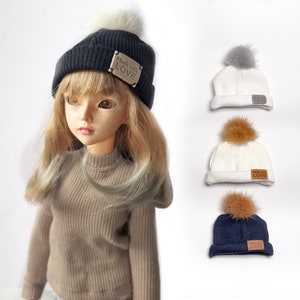 May include: Three knitted beanies with faux fur pom poms. Each beanie has a brown leather patch with the text "Made with LOVE". The beanies are in navy blue, white, and white with a brown pom pom.