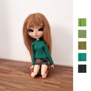 Könnte beinhalten: Eine Puppe mit langen braunen Haaren, die einen grünen Pullover und einen karierten Rock trägt. Die Puppe sitzt auf einem Holzboden.