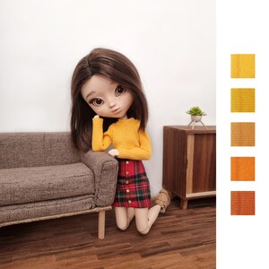 May include: A doll with brown hair wearing a yellow sweater and a red and black plaid skirt. The doll is kneeling on a wooden floor in front of a brown couch.