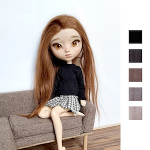 May include: A doll with long red hair wearing a black sweater and a plaid skirt, sitting on a brown sofa.