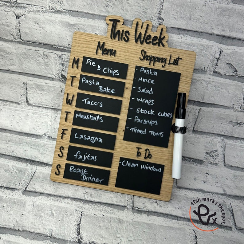 May include: A wooden weekly meal planner with a chalkboard surface. The planner has a section for "This Week", "Menu", and "Shopping List". The "Menu" section lists days of the week and meal ideas. The "Shopping List" section has a list of grocery items. The "To Do" section has a space to write tasks. The planner is made by "etch marks the spot".