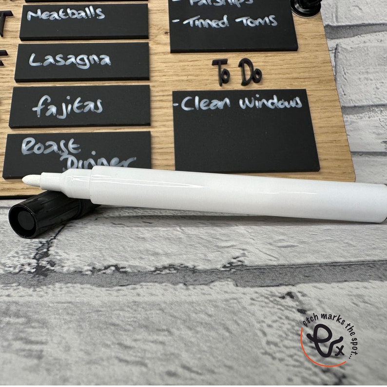 May include: A white dry-erase marker with a black cap, lying on a wooden surface with black chalkboard labels. The labels have handwritten text, including "Meatballs", "Lasagna", "fajitas", "Roast Dinner", "Tinned Toms", "To Do", and "Clean Windows".