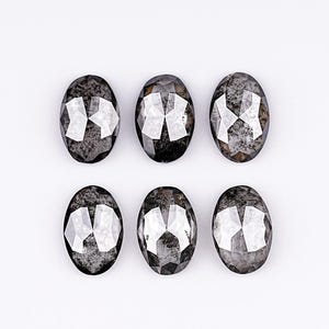 Op de afbeelding: Zes ovaalvormige, gefacetteerde edelstenen met een donkergrijze, gespikkelde uitstraling. De edelstenen zijn gerangschikt in twee rijen van drie op een witte achtergrond. Elke steen heeft een reflecterend, kristallijn interieur.