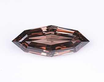 May include: A brown, elongated gemstone with multiple facets. The gem has a symmetrical, geometric shape, reflecting light. The gemstone is set against a plain white background, highlighting its colour and cut. The gem is likely for jewellery.