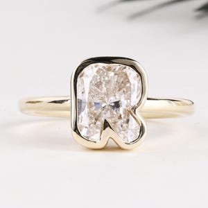 Puede incluir: Un anillo de oro con una piedra preciosa grande y clara en forma de letra. La piedra preciosa está engastada en un bisel y la banda es de oro pulido simple. El anillo se muestra sobre una superficie blanca, con una planta verde borrosa en el fondo.