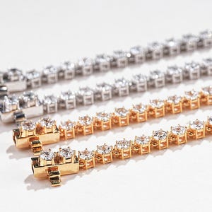K&ouml;nnte beinhalten: Drei Tennisarmb&auml;nder in Silber- und Goldt&ouml;nen, jedes mit zahlreichen runden, funkelnden Edelsteinen besetzt. Die Armb&auml;nder werden auf einer wei&szlig;en Oberfl&auml;che pr&auml;sentiert, um ihr aufwendiges Design und ihre Brillanz hervorzuheben. Das goldene Armband befindet sich im Vordergrund.