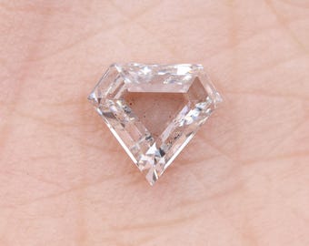 May include: A clear, faceted diamond with a unique geometric shape. The diamond has a triangular center and pointed edges, reflecting light. The gemstone is displayed on a light skin tone background.