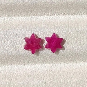 Può includere: Un paio di piccoli orecchini a lobo color rosa magenta vibrante a forma di stelle a sei punte. Gli orecchini presentano una superficie liscia e lucida e sono disposti fianco a fianco, mettendo in risalto la loro silhouette a stella e il colore saturo.