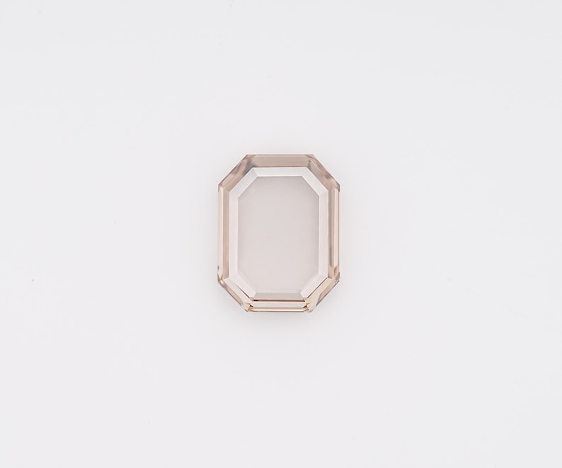 May include: A faceted, rectangular gemstone with a light brown hue. The gem has an emerald cut with a clear, rectangular center and a beveled edge. The gemstone is set against a white background.