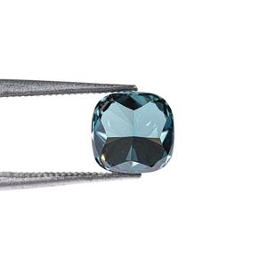 May include: A faceted, cushion-cut gemstone in shades of teal and blue is held by a pair of metal tweezers against a white background. The gemstone's facets reflect light, creating a sparkling effect. The overall presentation is clean and professional.