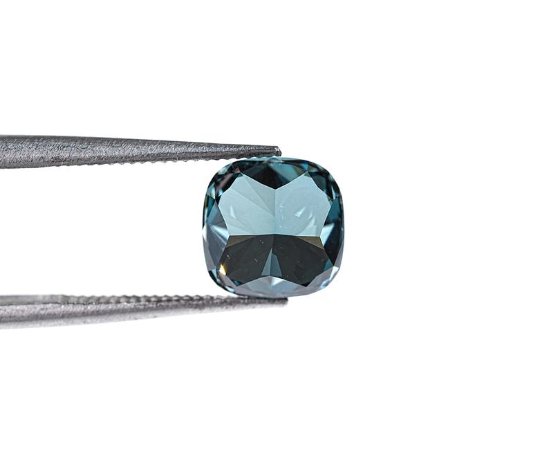 May include: A faceted, cushion-cut gemstone in shades of teal and blue is held by a pair of metal tweezers against a white background. The gemstone's facets reflect light, creating a sparkling effect. The overall presentation is clean and professional.