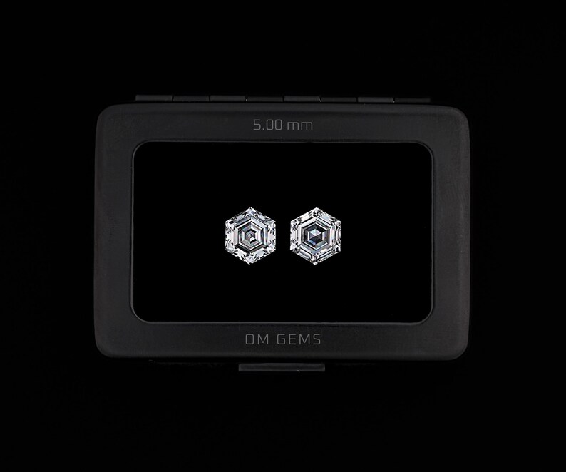 Puede incluir: Dos piedras preciosas transparentes de corte hexagonal exhibidas dentro de un estuche rectangular negro. El estuche presenta el texto OM GEMS y una medida de 5,00 mm impresa en el marco. Las piedras tienen una superficie facetada.