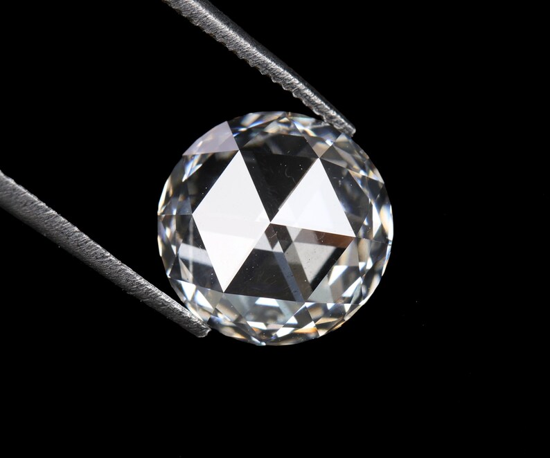 May include: A clear, round-cut diamond held by silver tweezers against a black background. The diamond's facets reflect light, creating a brilliant sparkle. The gem is a focal point, showcasing its clarity and cut.