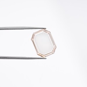 May include: A faceted, rectangular gemstone with a light peach-colored frame and a white interior. The gem is held by a pair of silver tweezers against a white background. The gem's shape is an octagon with a rectangular center.
