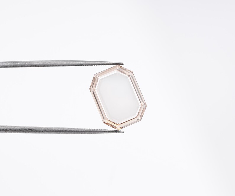 May include: A faceted, rectangular gemstone with a light peach-colored frame and a white interior. The gem is held by a pair of silver tweezers against a white background. The gem's shape is an octagon with a rectangular center.