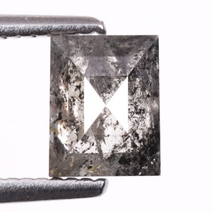 Puede incluir: Un diamante de forma cuadrada, gris, con un aspecto nublado y moteado negro. El diamante está sujeto por un par de pinzas metálicas.