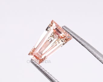 May include: A peach-colored, baguette-cut gemstone held by silver tweezers. The gem has a rectangular shape with angled facets, reflecting light. The image is set against a white background. The text "ONGEMINDIA" is visible on the lower left.