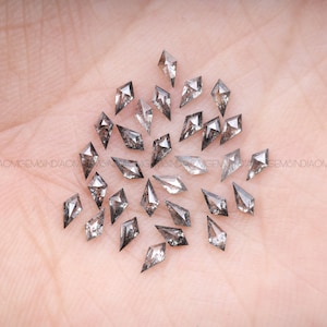 Puede incluir: Una colección de piedras preciosas grises facetadas en forma de diamante, también conocidas como diamantes en bruto, sobre una superficie rosa claro.