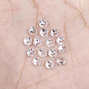 Puede incluir: Una colecci&oacute;n de catorce peque&ntilde;as piedras preciosas transparentes de corte hexagonal con superficies facetadas. Cada piedra presenta una forma geom&eacute;trica de seis lados con una parte superior puntiaguda, reflejando la luz a trav&eacute;s de sus m&uacute;ltiples facetas pulidas, dispuestas en un grupo suelto.